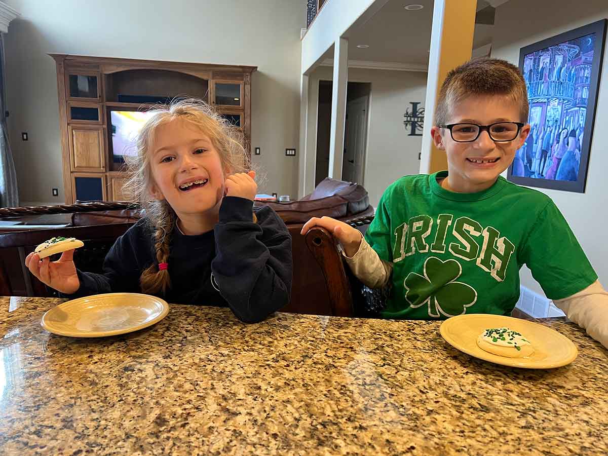 Girl in pigtail braids and boy in a green "Irish" shirt eating sugar cookies at a kitchen counter.
