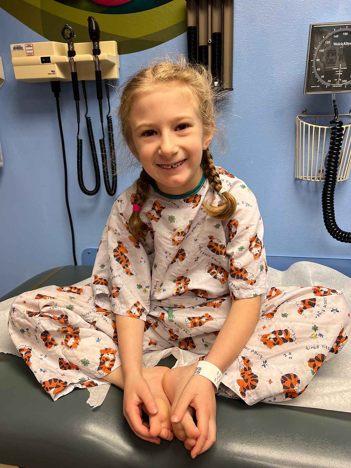 Little girl in French braid pigtails sitting on an exam table in hospital gown.