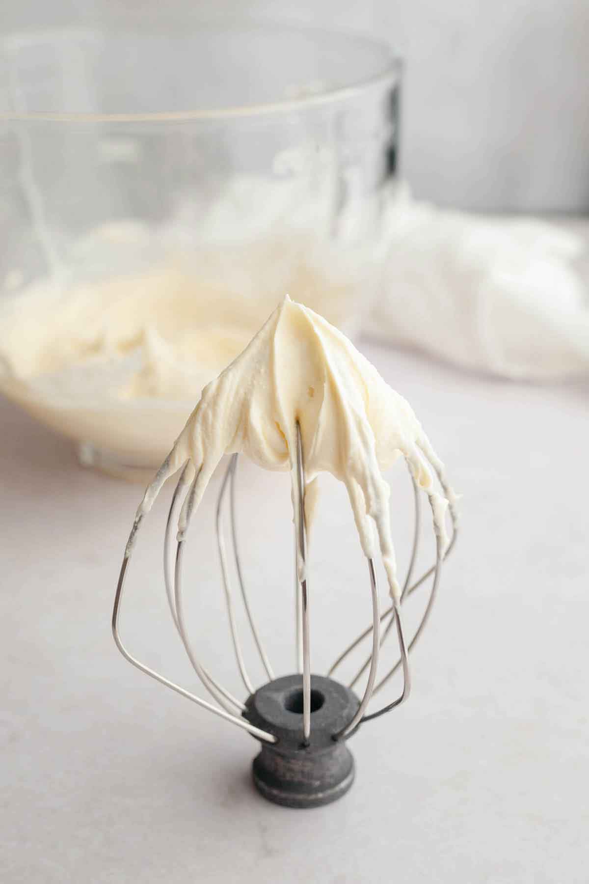 Wire whisk of a stand mixer sitting upright on a counter with homemade whipped cream in stiff peaks on the end.