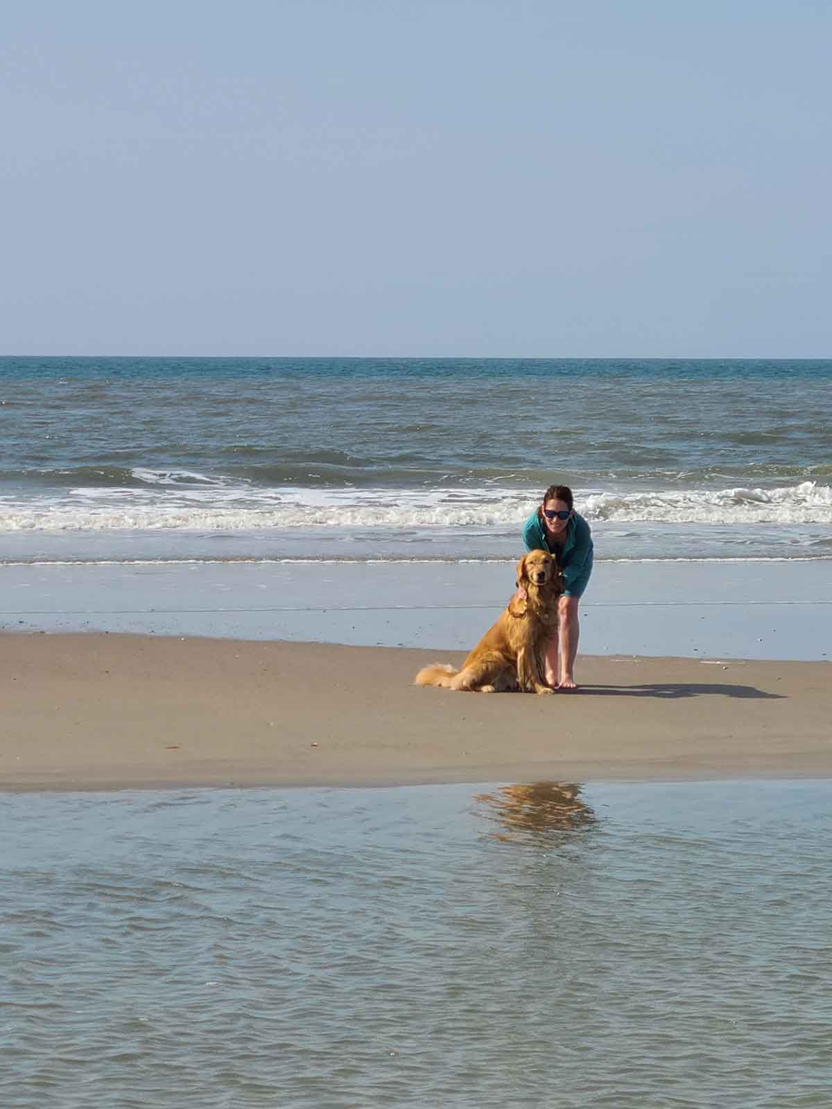 Woman and a golden retriever standing on a sand bar at the beach.