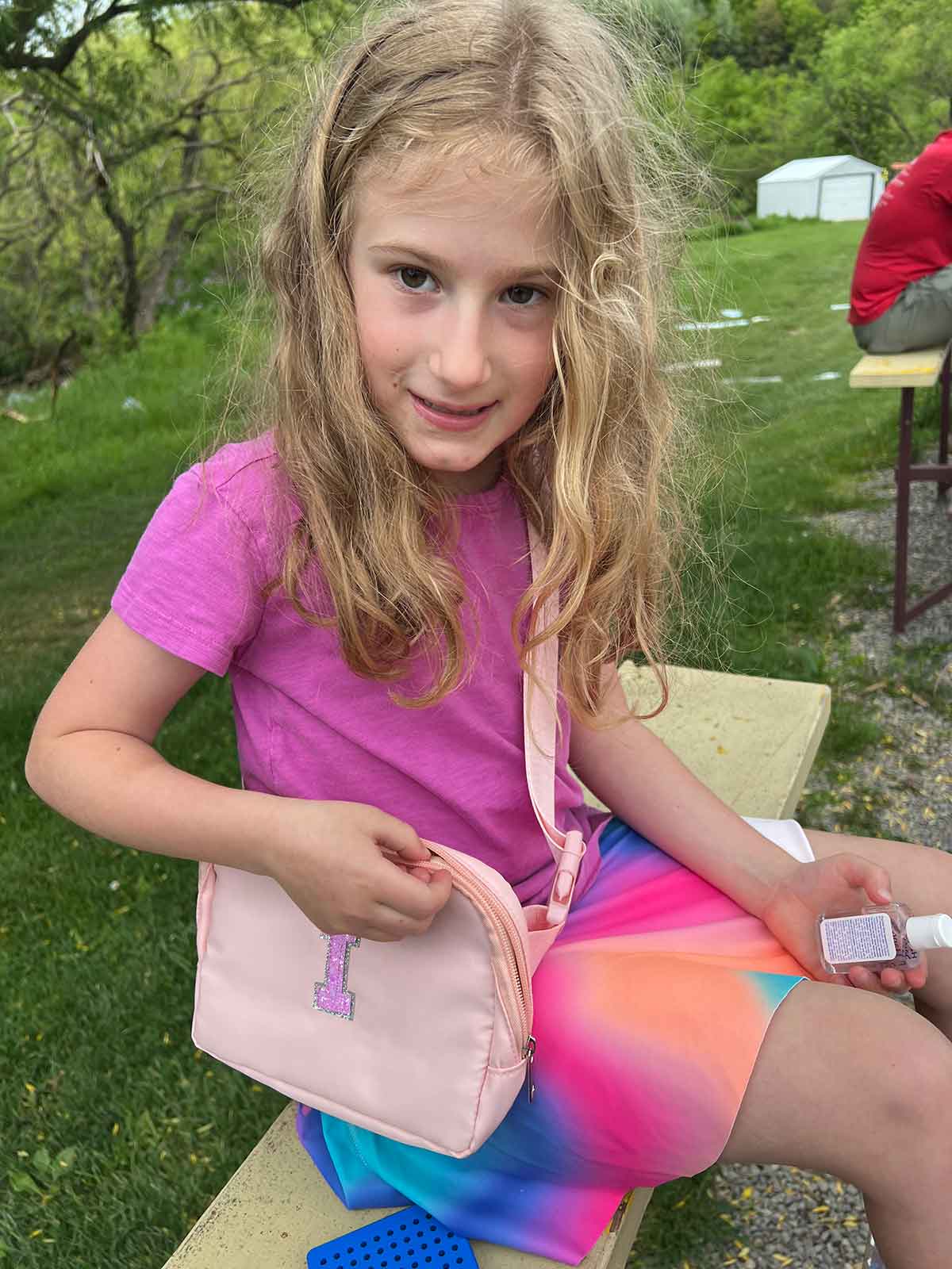 Little girl sitting on wooden bleachers with woods behind her, a cross body purse across her shoulder.