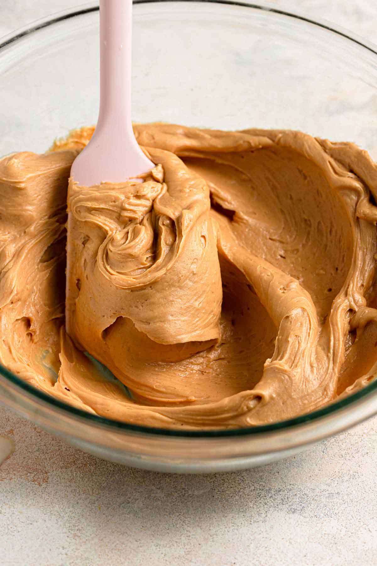 A pink spatula stirring peanut butter frosting in a glass bowl.