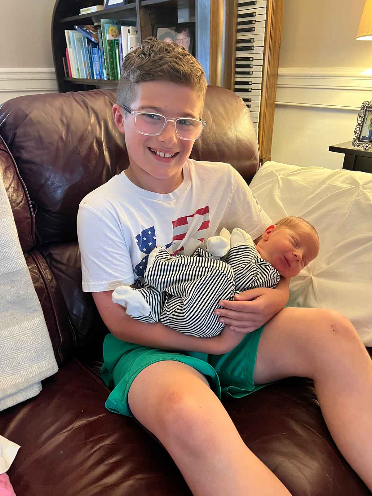 Ten year old boy in shorts and a tshirt sitting on a couch holding a newborn baby.