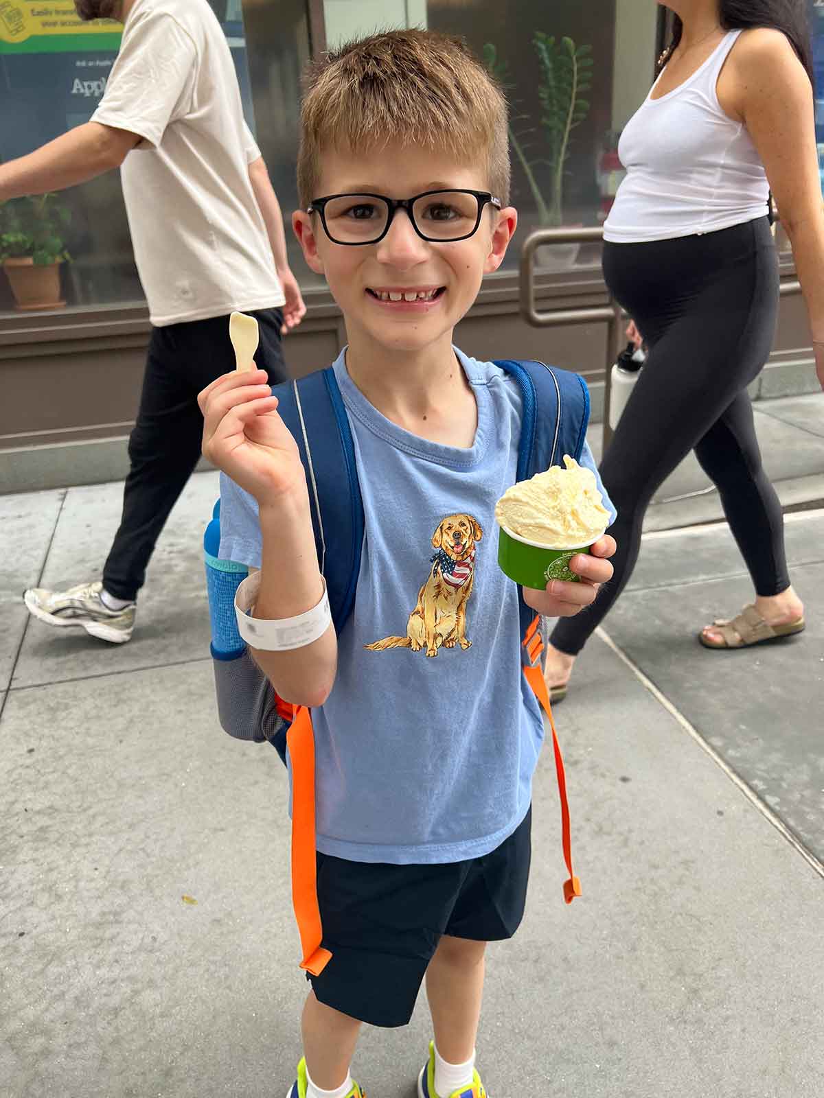 Little boy standing on a city street eating gelato out of a cup and smiling.