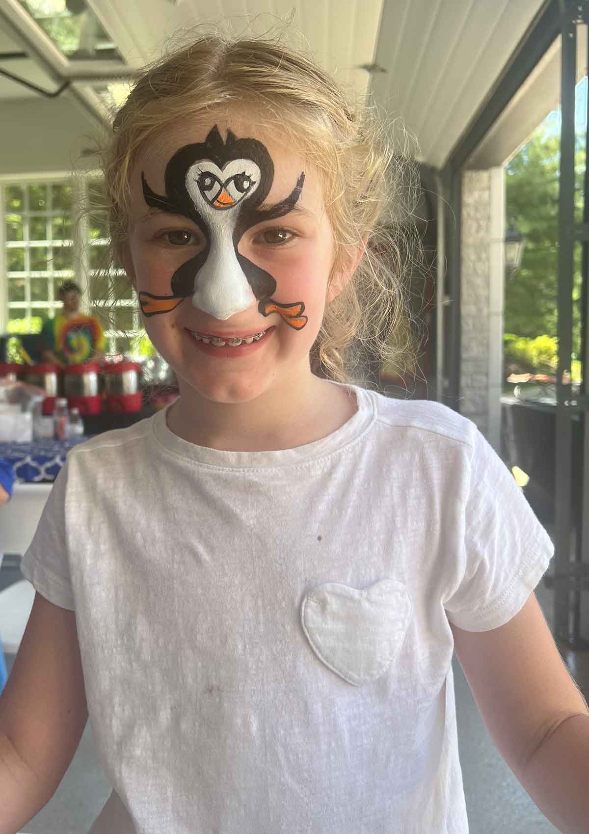 Little girl wearing a white tshirt with her face painted as a penguin.