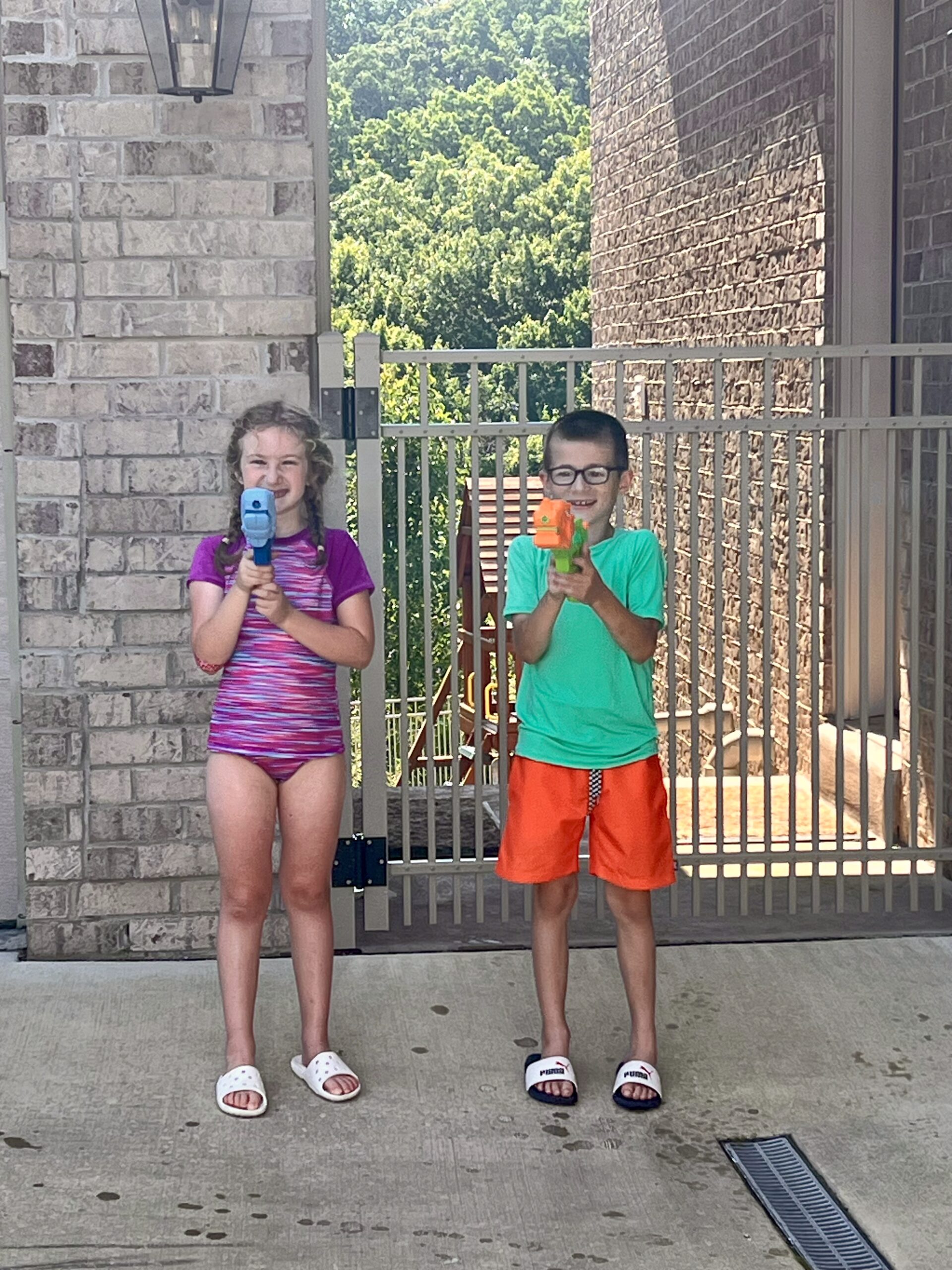 A girl and a boy in their swimsuits playing with water guns in a driveway.