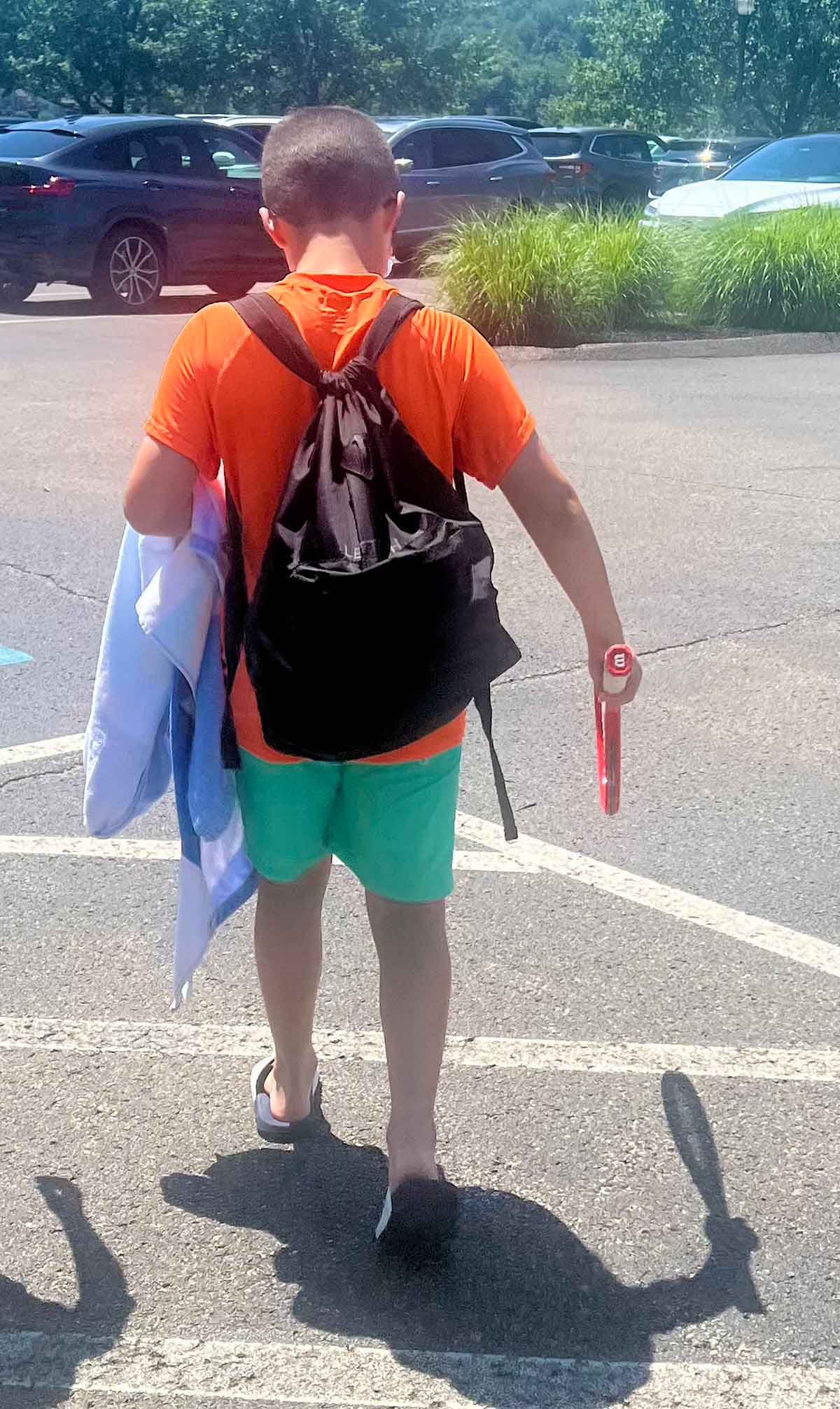 Photo of a boy from behind walking through a parking lot in a swimsuit, carrying a towel and a tennis racket.