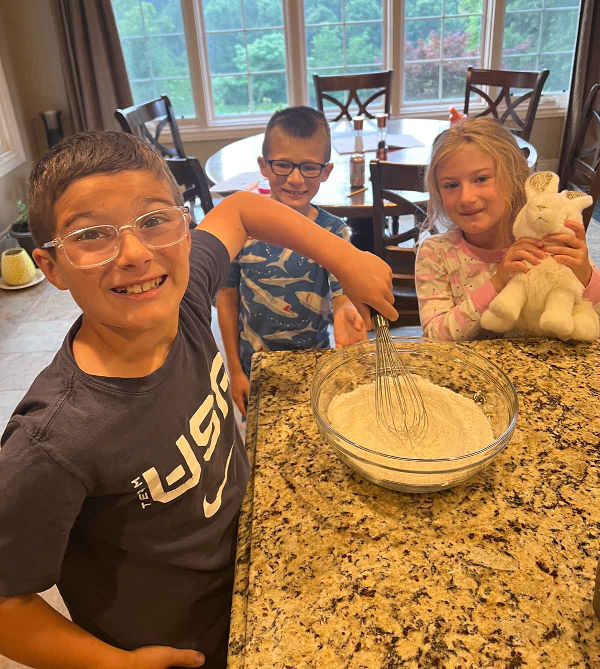 Three kids standing at a kitchen counter whisking a bowl of pancake batter.