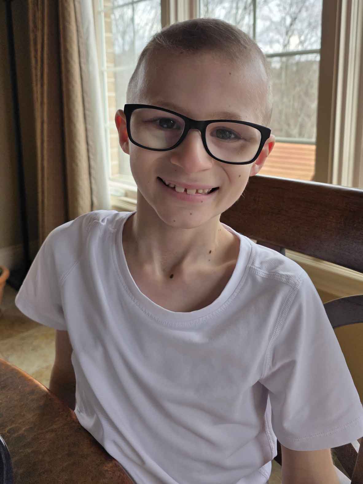 Little boy in glasses sitting at a kitchen table in front of windows.