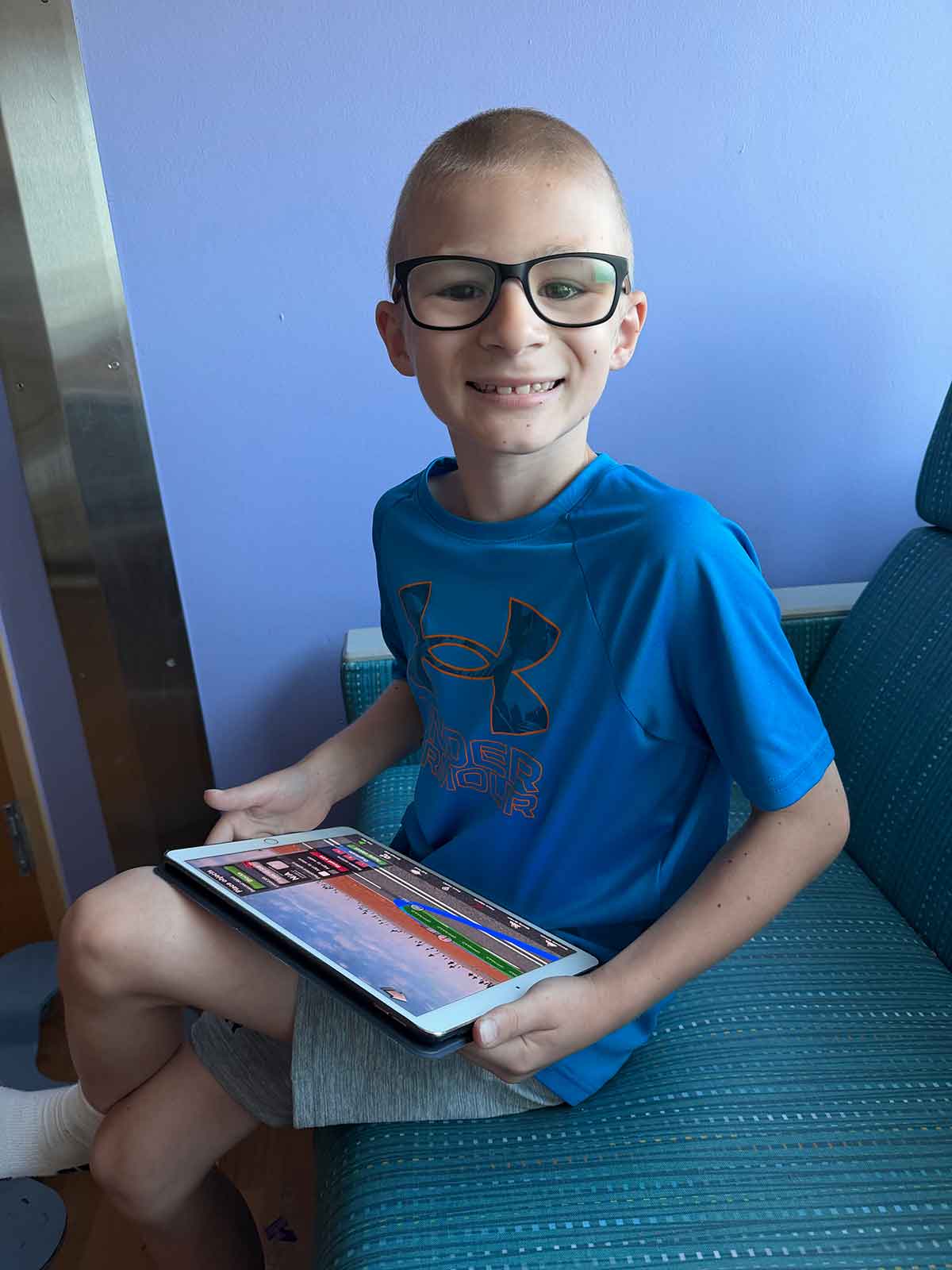 Boy in a blue shirt and glasses looking up from an ipad smiling.